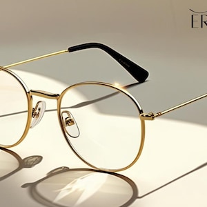 May include: Gold-rimmed eyeglasses with a round frame and black temples. The glasses are set against a neutral background, casting a shadow. The brand name "ERATIQUE EYEWEAR" is displayed in the upper right corner.