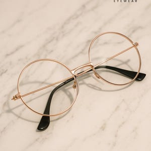 May include: Round eyeglasses with a rose gold frame and black temples. The glasses have clear lenses and are displayed on a marble surface. The brand name "ERATIQUE EYEWEAR" is visible in the upper right corner.