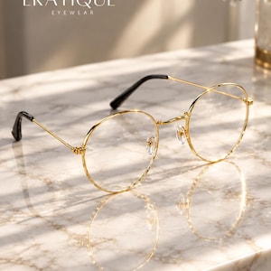 May include: Gold-toned round eyeglasses with thin metal frames, clear lenses, and black temple tips. The brand name ERATIQUÉ EYEWEAR is printed in white text above the glasses. The design features adjustable nose pads.