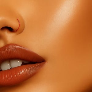 May include: Close-up of a person's face featuring a red nose ring. The image highlights the lips with a glossy, brown lipstick. The skin has a warm, orange tone. The brand name "ERATIQUE" is at the bottom right.