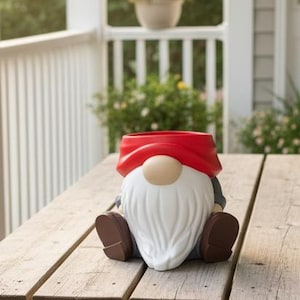May include: A gnome-shaped planter featuring a bright red hat, a large beige nose, and a long white beard. The gnome is depicted in a seated position with brown boots and dark gray sleeves, with an open top designed for holding small plants.