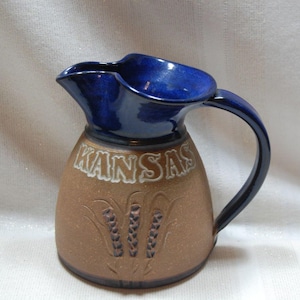 May include: A ceramic pitcher with a brown base and a deep blue top and handle. The word "KANSAS" is displayed in white lettering on the front, with wheat stalks below. The pitcher has a curved handle and a spout for pouring.