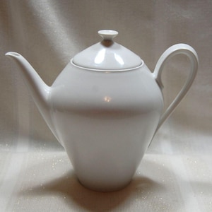 May include: A white ceramic teapot with a lid and a curved handle. The teapot has a long spout and a rounded body. The lid has a small knob on top. The teapot is on a white surface.