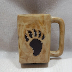 May include: A tan ceramic mug with a rectangular shape and a large, dark brown bear paw print. The mug has a matching handle and a marbled effect throughout the surface.