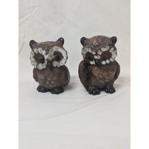 May include: Two decorative owl figurines. The owls are brown with white and brown accents around the eyes. They have black feet and ears. The figurines are made of a textured material and sit on a white surface.