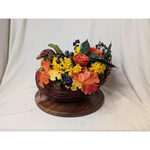 May include: A woven brown turkey-shaped basket filled with colorful artificial flowers and fall foliage. The arrangement includes yellow, orange, and purple flowers, with decorative leaves. The basket sits on a round wooden base.