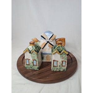May include: A ceramic windmill-shaped container with a blue and white design, flanked by two house-shaped containers. The containers have green and yellow roofs, and are set on a round wooden tray.
