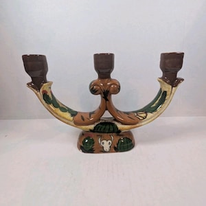 May include: A ceramic candelabra with three candle holders, featuring a curved base decorated with hand-painted green cacti and a white bull skull motif. The piece is finished in shades of brown, cream, and green with a glossy glaze throughout.