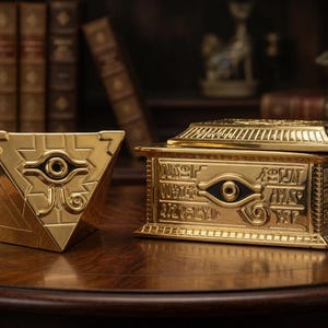 Könnte beinhalten: Zwei goldfarbene Sammlerboxen auf einem Holztisch. Eine ist pyramidenförmig, die andere eine rechteckige Box mit Deckel. Beide haben ein Augendesign und gravierte Hieroglyphen, vor dem Hintergrund antiker Bücher.