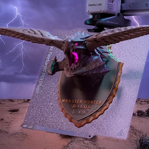 Op de afbeelding: Een 3D-geprinte drakenkop-trofee met uitgestrekte vleugels, gemonteerd op een schildvormige plaat. De plaat bevat de tekst "MONSTER HUNTER WILDS". De achtergrond toont een woestijnlandschap met bliksem.