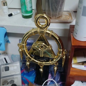 May include: A gold-colored, circular pendant with a central triangle featuring an eye design. The pendant has a ring at the top for hanging and decorative spikes hanging from the bottom. The pendant is likely a decorative item or accessory.