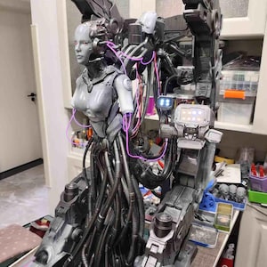 May include: A detailed gray cyborg figure with red eyes, integrated into a large mechanical structure with dark metallic components. Bright pink cables connect the figure to the machinery, which features a small screen displaying glowing icons.