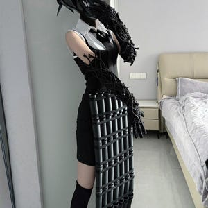 Könnte beinhalten: Eine Person in einem schwarzen Kostüm mit Helm, Handschuhen und einem schwarzen Kleid. Das Kostüm beinhaltet ein schwarzes, rechteckiges Objekt mit mehreren zylindrischen Röhren. Das Outfit wird mit schwarzen kniehohen Socken und Schuhen ergänzt.