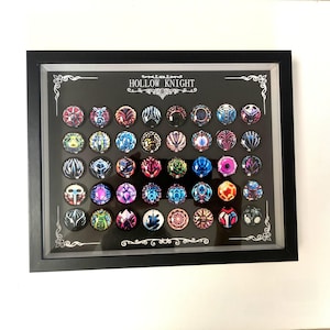 May include: A framed display of circular pins with detailed designs in various colours. The pins are set against a black background, with the text "HOLLOW KNIGHT" above. The black frame has decorative corner detailing.