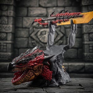 Op de afbeelding: Gedetailleerd beeldje met een rode en zwarte drakenkop die een groot zwaard met een geel lemmet ondersteunt. Het zwaard heeft rode accenten en wordt omhoog gehouden door een grijze, getextureerde basis. De bek van de draak is open en toont witte tanden.