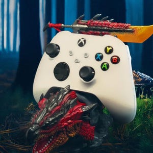 Könnte beinhalten: Ein weißer Xbox-Controller mit schwarzen Tasten und farbigen Aktionstasten, der auf einem roten und schwarzen Drachenkopf-Sockel ruht. Ein dekoratives Schwert mit roten und gelben Akzenten sitzt auf dem Controller und erzeugt ein Fantasy-Gaming-Thema.