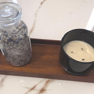 Puede incluir: Una bandeja de madera contiene un tarro de cristal transparente lleno de sales de baño y una vela negra en un recipiente de cristal redondo. La vela tiene tres mechas y cera blanca. El tarro tiene una tapa de cristal transparente.