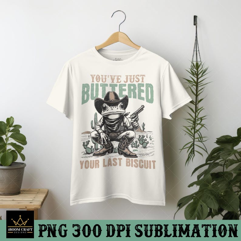 K&ouml;nnte beinhalten: Wei&szlig;es T-Shirt mit einem Grafikdruck einer Frosch, der einen Cowboyhut tr&auml;gt und eine Pistole h&auml;lt. Der Text "You've Just Buttered Your Last Biscuit" ist auf dem T-Shirt aufgedruckt.