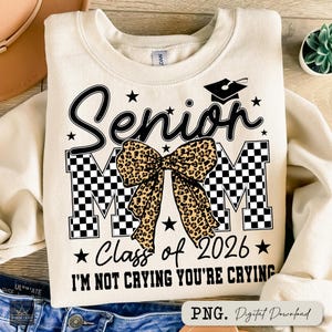 May include: Cream-colored sweatshirt with the text "Senior Mom" in black script and block letters, featuring a leopard print bow. Includes "Class of 2026" and the phrase "I'M NOT CRYING YOU'RE CRYING".