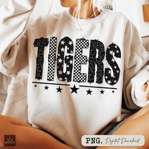 May include: Cream-colored sweatshirt with the word "TIGERS" in a bold, multi-patterned design. The letters feature leopard print, checkered patterns, and star-filled designs. Black stars are aligned below the word, with the text "PNG. Digital Download" in the bottom right corner.