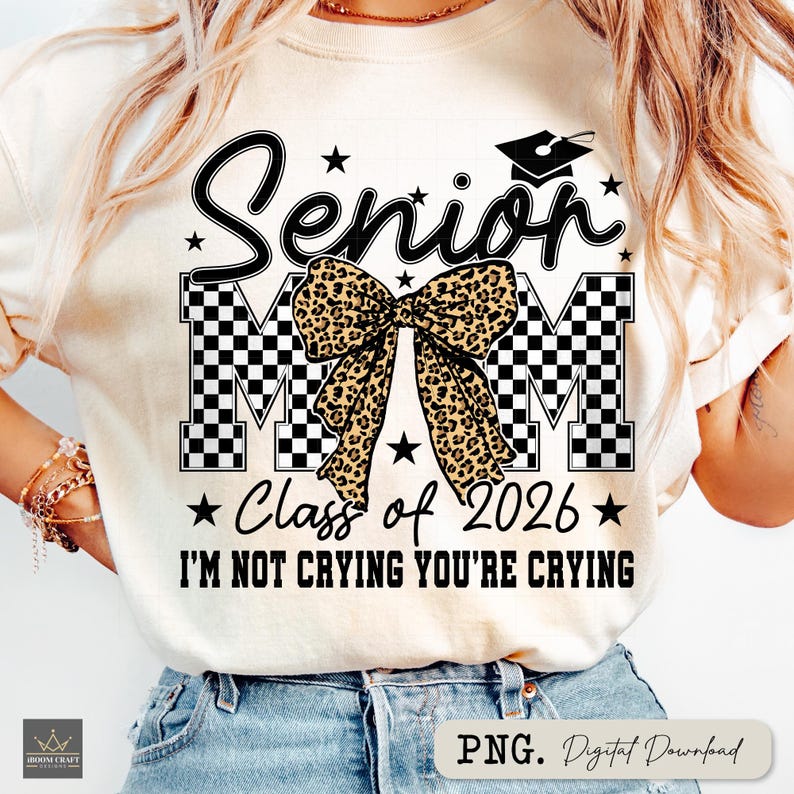 May include: Cream-colored t-shirt with a black and white checkered design spelling out "MOM" and the word "Senior" in script. A leopard print bow is centered above the "MOM" letters. The text "Class of 2026" and "I'M NOT CRYING YOU'RE CRYING" are also printed on the shirt.