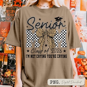 May include: A tan t-shirt with a graphic design for a Senior Mom. The design includes the word "Senior" in black script, the letters "MOM" in a checkered pattern, a leopard print bow, and the text "Class of 2026". The phrase "I'M NOT CRYING YOU'RE CRYING" is also included.