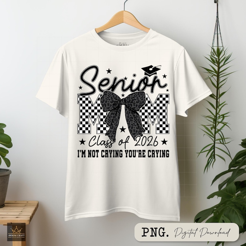 May include: White t-shirt with a graduation theme. The shirt features the word "Senior" in a script font, with "MM" in a checkered pattern. A bow with a leopard print design is in the center. The text "Class of 2026" and "I'M NOT CRYING YOU'RE CRYING" are also included.