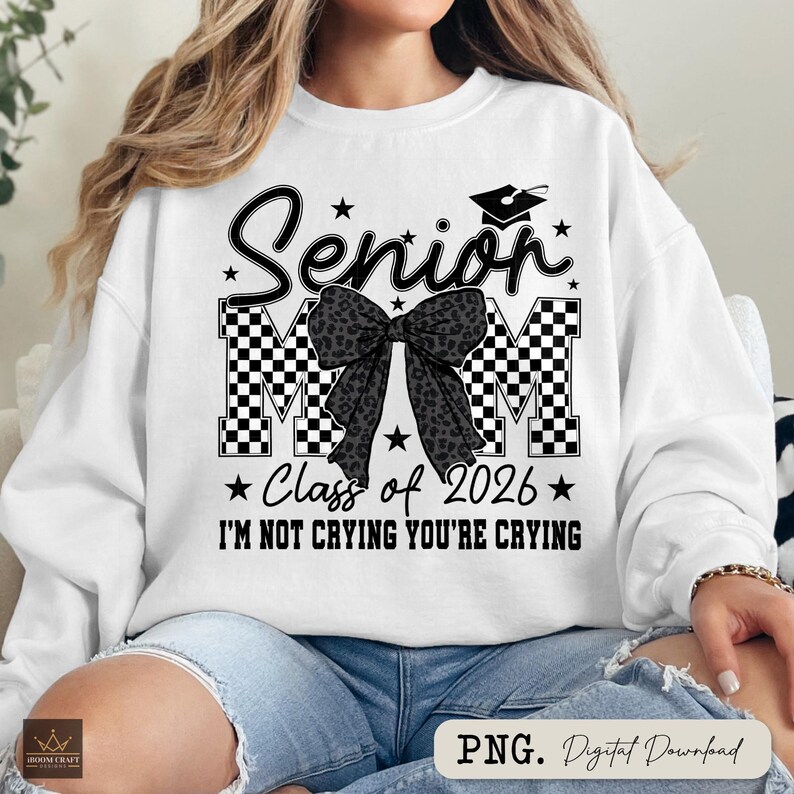 May include: White sweatshirt with a black and white design. The design includes the word "Senior", a graduation cap, stars, and the text "Class of 2026" and "I'M NOT CRYING YOU'RE CRYING". A black bow with a leopard print pattern is in the center.