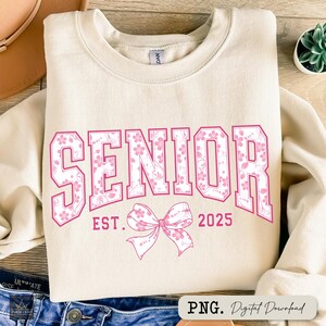 Pu&ograve; includere: Felpa color crema con la scritta "SENIOR" in rosa con stampa floreale. Sotto, le parole "EST. 2025" e un fiocco rosa. La felpa &egrave; piegata sopra dei jeans.