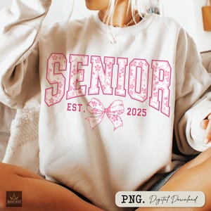 Pu&ograve; includere: Una felpa color crema con la scritta "SENIOR" in grandi lettere rosa a motivi floreali. Sotto, un fiocco rosa e il testo "EST. 2025". La felpa &egrave; in stile casual, ideale per le celebrazioni di laurea.