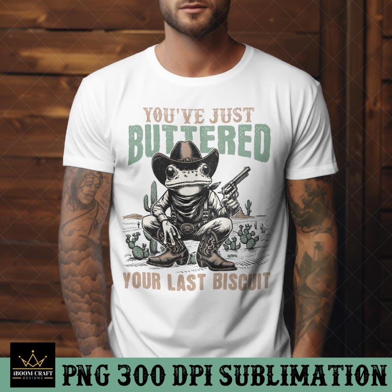 K&ouml;nnte beinhalten: Wei&szlig;es T-Shirt mit einem Grafikdruck einer Frosch, der einen Cowboyhut tr&auml;gt und eine Pistole h&auml;lt. Der Text "You've Just Buttered Your Last Biscuit" ist unter dem Frosch gedruckt.