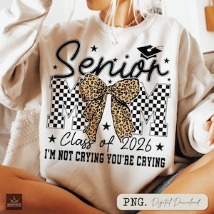 May include: Cream-colored sweatshirt with the text "Senior Mom" in black script, a graduation cap, and the words "Class of 2026." A leopard print bow is centered on the design, with the phrase "I'm not crying you're crying" below.