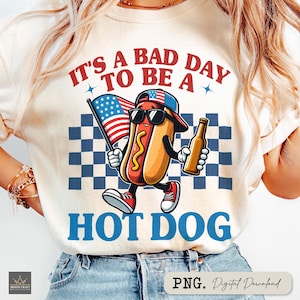 May include: Cream-colored t-shirt with a cartoon hot dog wearing sunglasses and a baseball cap, holding an American flag and a beer bottle. The text reads "IT'S A BAD DAY TO BE A HOT DOG" in red and blue.