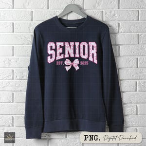 Pu&ograve; includere: Felpa blu navy con la scritta "SENIOR" in lettere floreali rosa. Sono presenti anche il testo "EST. 2025" e un fiocco rosa. La felpa &egrave; appesa a una gruccia grigia contro un muro di mattoni bianchi.
