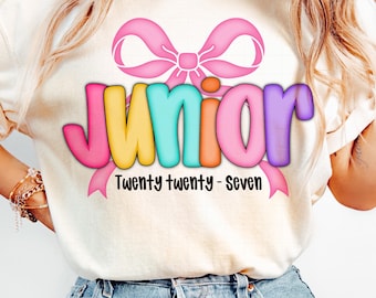 Könnte beinhalten: Cremefarbenes T-Shirt mit einem bunten "Junior"-Motiv. Das Wort "Junior" ist in Pink, Gelb, Türkis, Orange und Lila gehalten, mit einer rosa Schleife darüber. Darunter steht der Text "Twenty twenty - Seven".