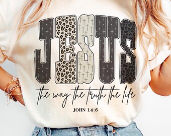 May include: Cream-colored t-shirt with the word "JESUS" in large, bold letters. The letters have different patterns, including leopard print and crosses. Below the word are the words "the way the truth the life" and the text "JOHN 14:6".