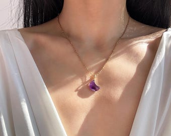 May include: A delicate gold chain necklace featuring a small, polished purple amethyst gemstone carved into a crescent moon shape. The pendant hangs from a simple gold bail, suspended on a thin, textured gold link chain that rests against the collarbone.