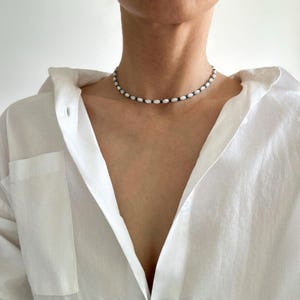 Peut inclure: Un collier ras-du-cou composé d'un motif répétitif de petites perles blanches de forme ovale, alternées avec des perles bleu foncé plus petites. Le collier est porté près du cou, contrastant avec une chemise blanche à col simple et épurée.