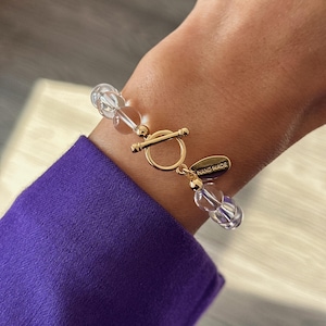May include: A bracelet featuring clear, spherical beads and a gold-toned toggle clasp. The clasp includes a small, oval-shaped gold-toned charm with the text HAND MADE engraved on its surface, connecting the two ends of the beaded bracelet.