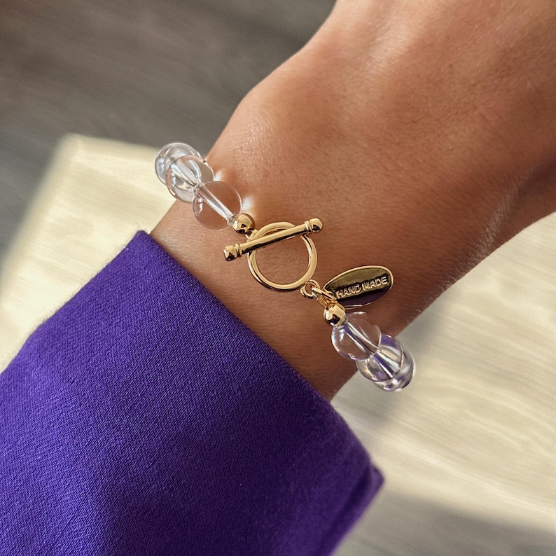 May include: A bracelet featuring clear, spherical beads and a gold-toned toggle clasp. The clasp includes a small, oval-shaped gold-toned charm with the text HAND MADE engraved on its surface, connecting the two ends of the beaded bracelet.