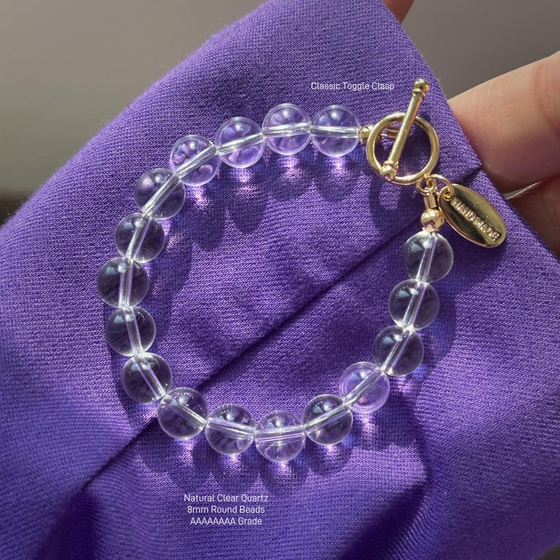 May include: A bracelet featuring 8mm round natural clear quartz beads with a gold-toned classic toggle clasp and a small oval tag engraved with the word HANDMADE. The clear beads are arranged in a circular shape on a vibrant purple fabric.