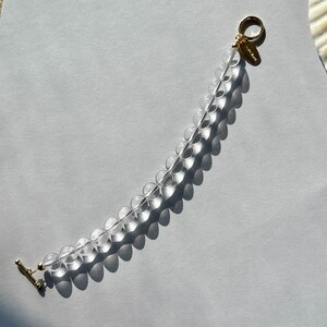 May include: A bracelet featuring a single strand of clear, spherical beads. The piece is finished with a gold-toned toggle clasp and a small, oval-shaped gold-toned metal tag attached near the closure, creating a simple and elegant design.