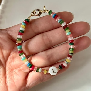Peut inclure: Un bracelet en perles colorées avec un charme blanc orné de la lettre "L" en lettres dorées. Le bracelet est composé de petites perles rondes de différentes couleurs : rouge, vert, bleu, jaune et rose.