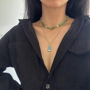May include: A layered necklace set featuring a gold chain with a rectangular pendant and a green beaded choker. The pendant has a blue and red design. The necklaces are worn over a black corduroy shirt.