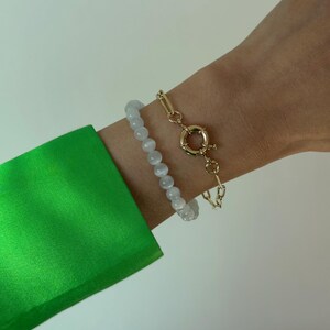 May include: A set of two bracelets consisting of one strand of smooth, translucent white spherical beads and one gold-toned metal chain bracelet featuring elongated oval links and a prominent circular spring ring clasp closure.