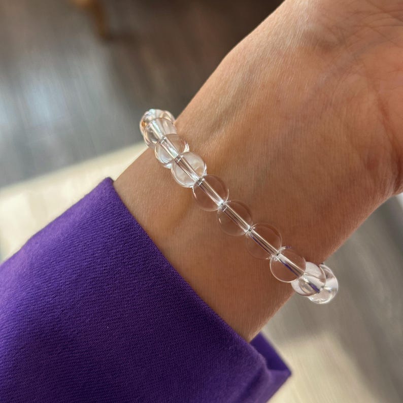 May include: A stretch bracelet composed of smooth, spherical, clear transparent beads. The beads are uniform in size and shape, strung together in a single continuous loop that encircles the wrist, reflecting light through the clear material.