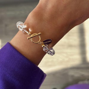 May include: A bracelet featuring clear, spherical beads strung together with a gold-toned toggle clasp. A small, oval-shaped purple charm hangs from the clasp, with the words HAND MADE clearly engraved in gold lettering on its surface.