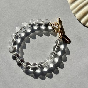 May include: A bracelet composed of clear, spherical beads strung together, featuring a gold-toned metal toggle clasp. A small, oval-shaped gold-toned metal tag is attached to the clasp, with the word HANDMADE clearly visible on its surface.
