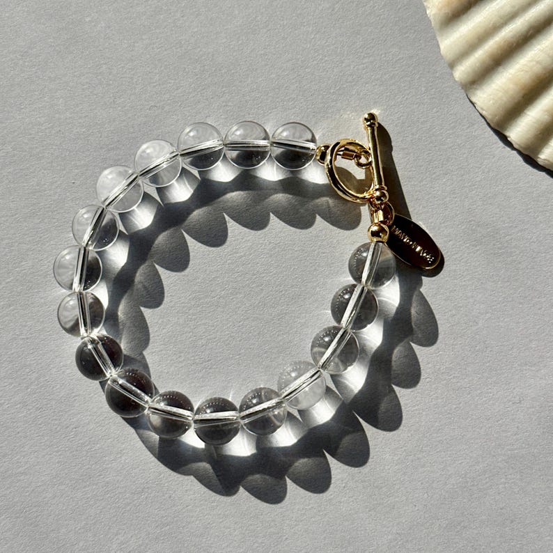 May include: A bracelet composed of clear, spherical beads strung together, featuring a gold-toned metal toggle clasp. A small, oval-shaped gold-toned metal tag is attached to the clasp, with the word HANDMADE clearly visible on its surface.