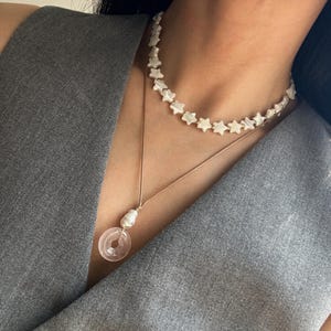 Puede incluir: Un collar de dos capas con una cadena delicada y un colgante con un círculo rosa con una perla. El collar superior está hecho de cuentas blancas en forma de estrella.